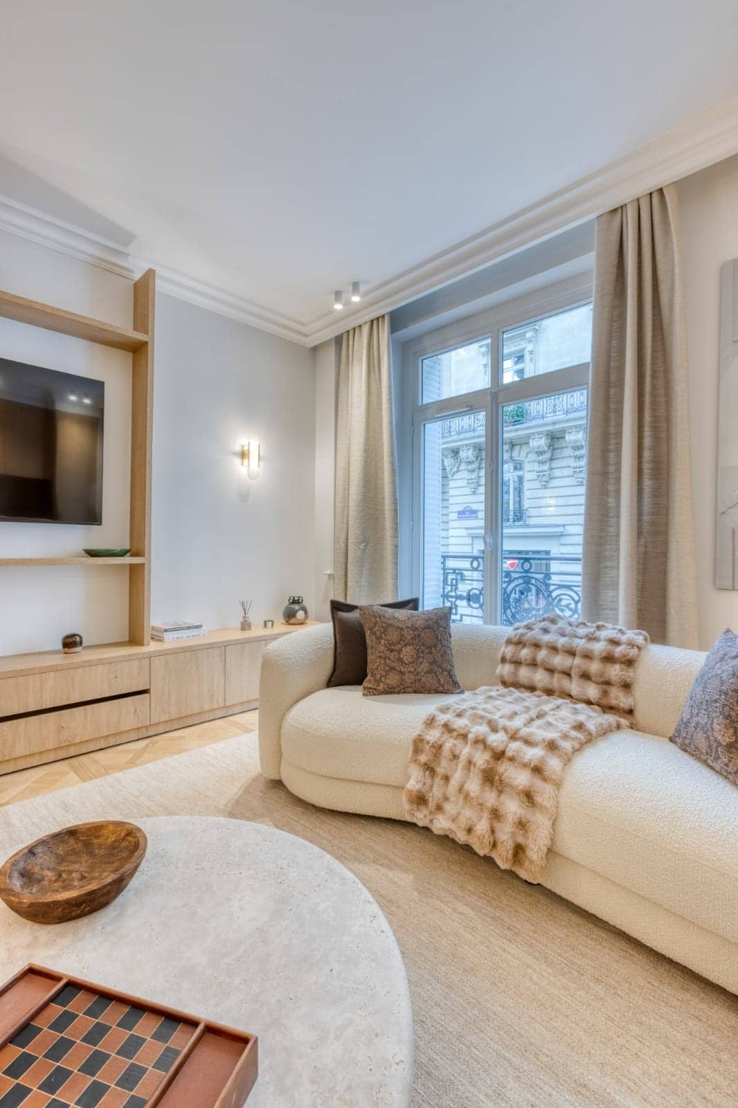 Salon avec meuble télévision en chêne, canapé en bouclé crème, rideaux beiges et fenêtre sur rue