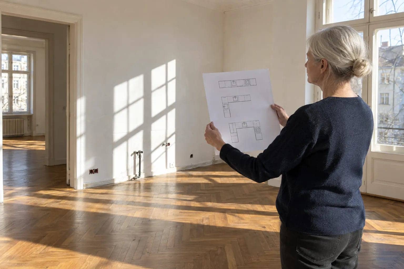 Une femme âgée tient un plan architectural devant une pièce vide avec parquet et grandes fenêtres.