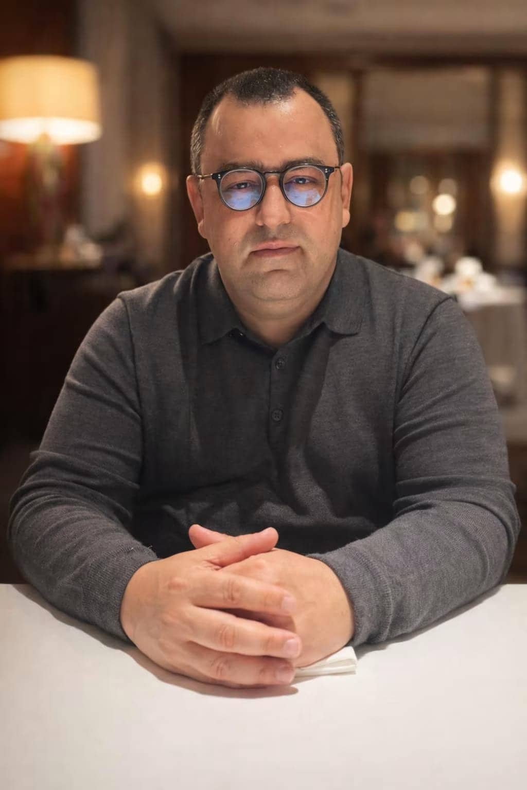Portrait d'Ilann Atlan, homme à lunettes en pull gris, assis les mains jointes