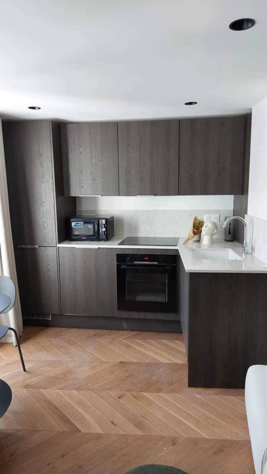 Cuisine d'angle en bois gris foncé, plan de travail clair, four noir et parquet en chevrons