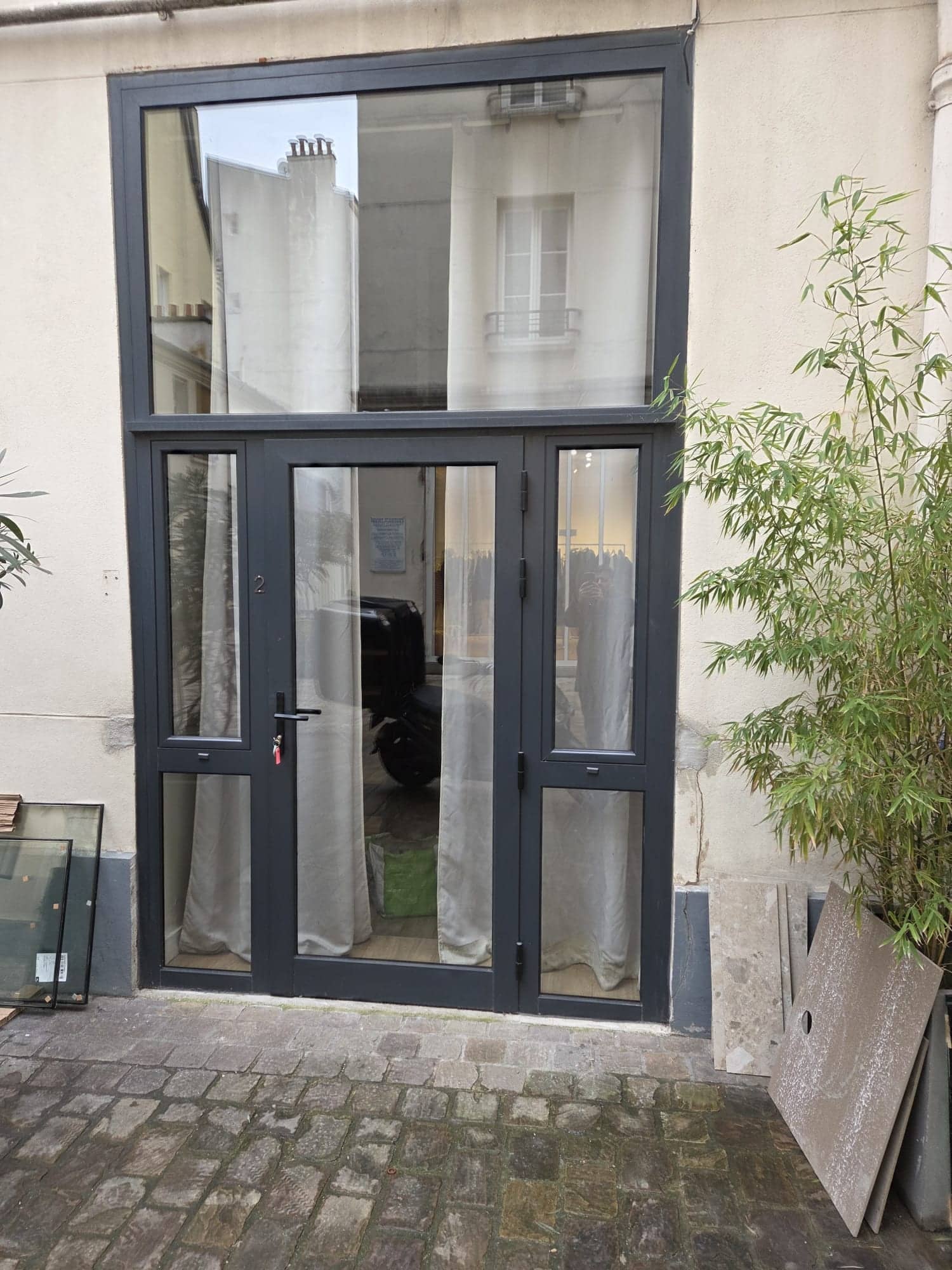 Entrée vitrée à menuiseries gris anthracite dans une cour pavée, avec vitrages posés contre le mur